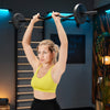 Woman in a yellow sports bra and black leggings lifting a barbell in a home gym setting.